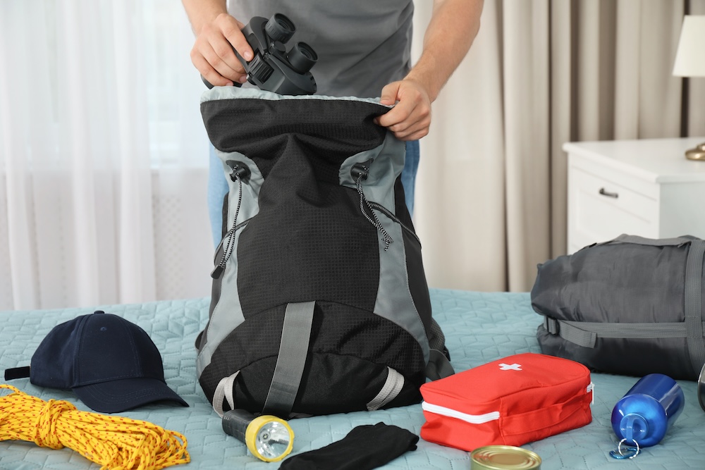 Man packing different camping equipment into backpack at home
